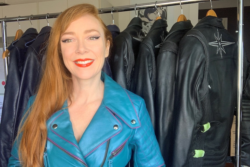 A woman with long red hair, wearing a bright blue leather jacket, stands in front of a rail of jackets.