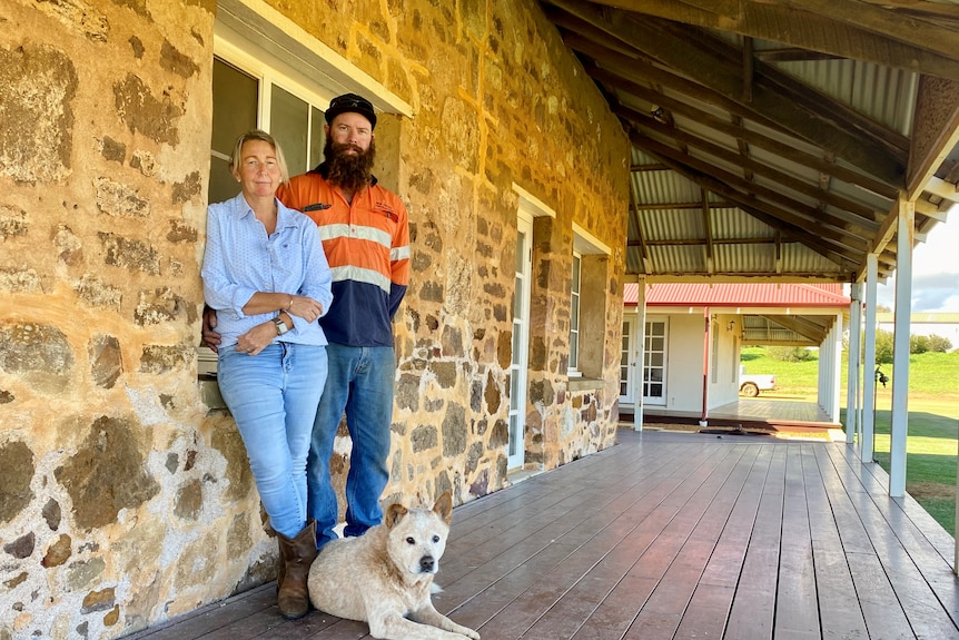 Lady in blue jeans and boots with man in beard and high vis shirt lean against stone wall with dog at their feet