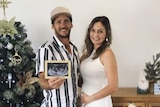 Couple stands holding ultrasound picture next to Christmas tree. Woman holds belly.
