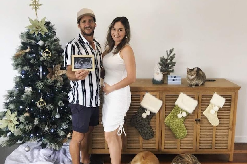 Couple stands holding ultrasound picture next to Christmas tree. Woman holds belly.