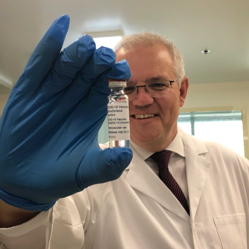 A man with grey hair and glasses holds up a vial of the AstraZeneca vaccine while wearing a white labcoat and blue gloves