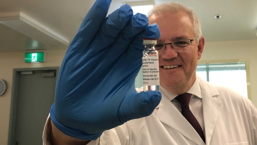 A man with grey hair and glasses holds up a vial of the AstraZeneca vaccine while wearing a white labcoat and blue gloves