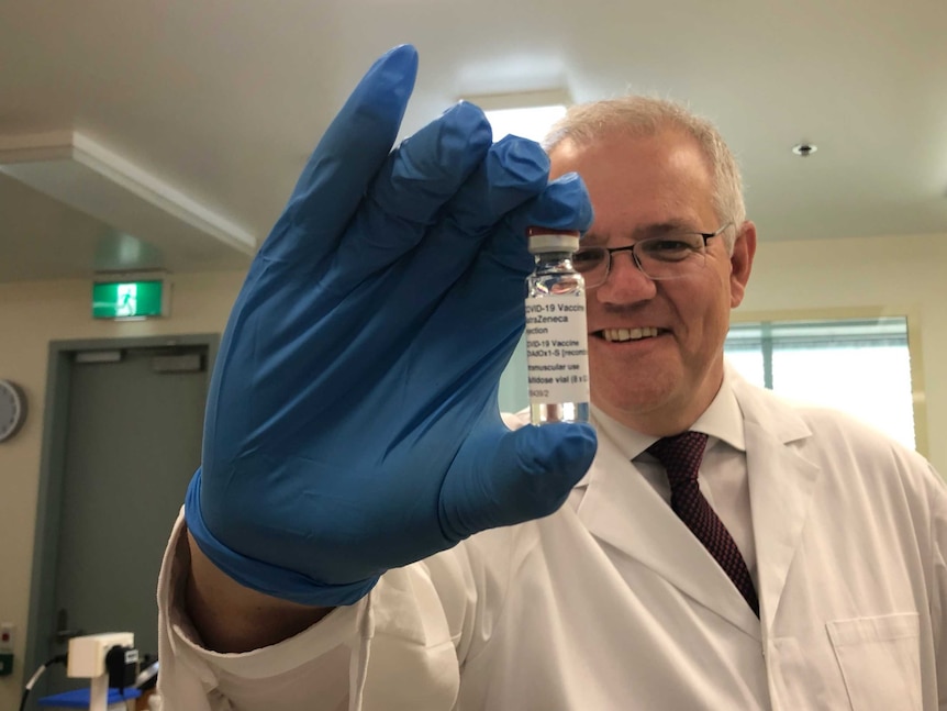 Un homme aux cheveux gris et aux lunettes tient un flacon de vaccin AstraZeneca tout en portant un sarrau blanc et des gants bleus