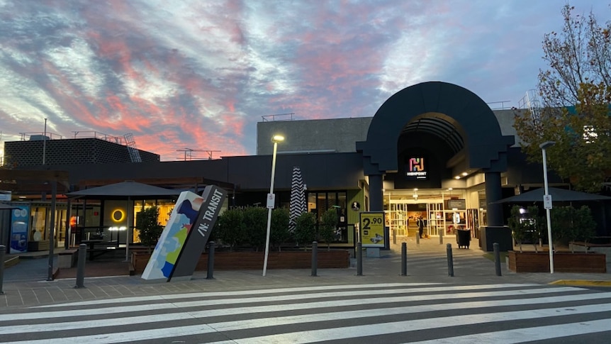Lights are on inside the Highpoint Shopping Centre, seen from outside under a red and blue morning sky.