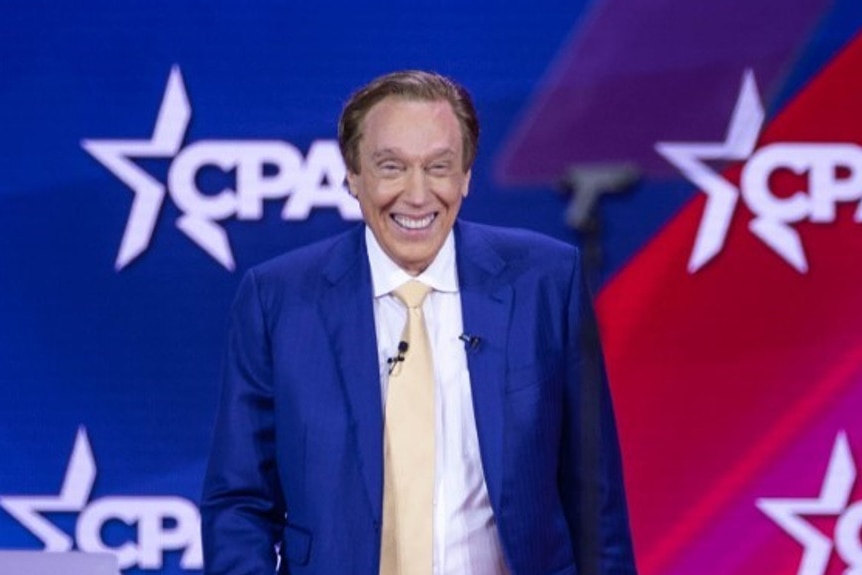 An older man with dyed hair in a blue suit walks onto the stage at CPAC.