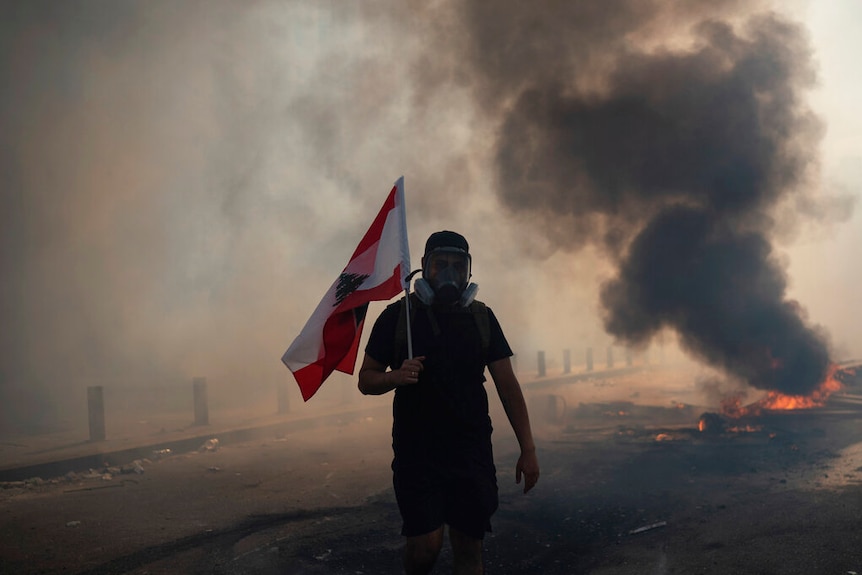 A protester wearing a gas mask, holding a Lebanese flag, walks n front of fire and smoke in the middle of street.