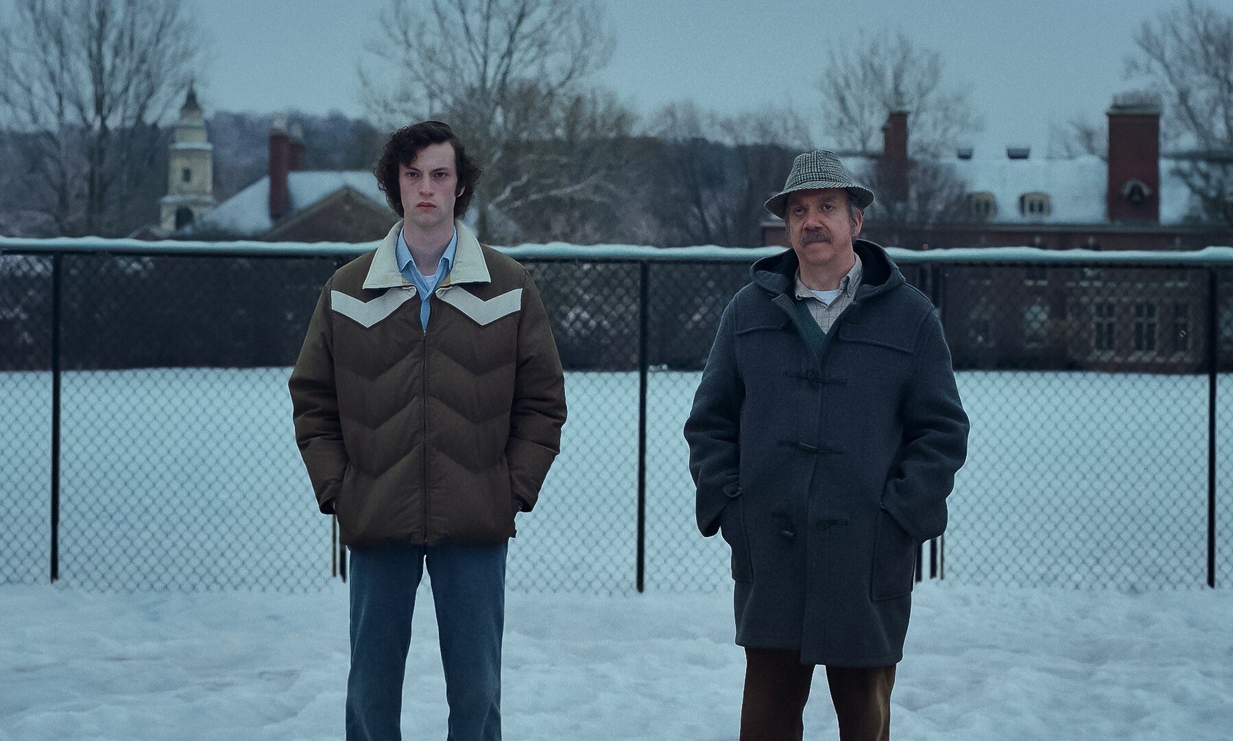 A film still of Dominic Sessa and Paul Giamatti standing outside on a snowy day, hands in pockets, with impassive expressions.