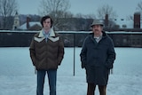 A film still of Dominic Sessa and Paul Giamatti standing outside on a snowy day, hands in pockets, with impassive expressions.