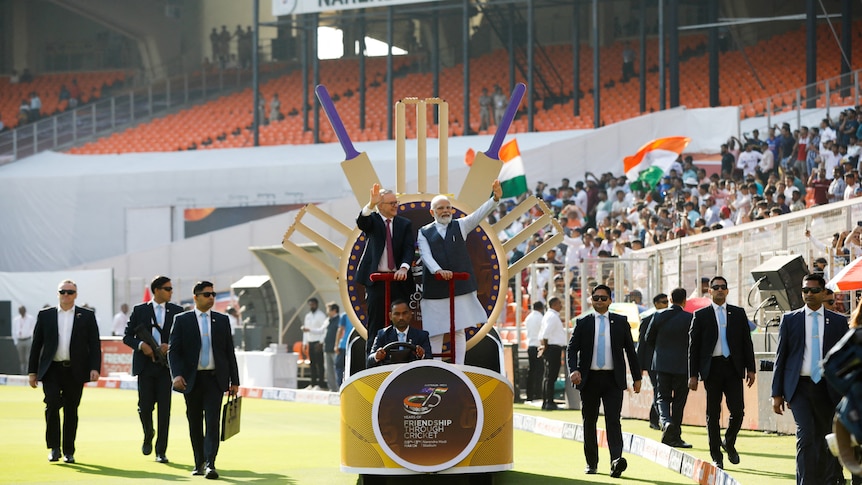 Anthony Albanese and Narendra Modi stand on a chariot with cricket bats on a cricket pitch surroudned by security.