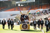 Anthony Albanese and Narendra Modi stand on a chariot with cricket bats on a cricket pitch surroudned by security.