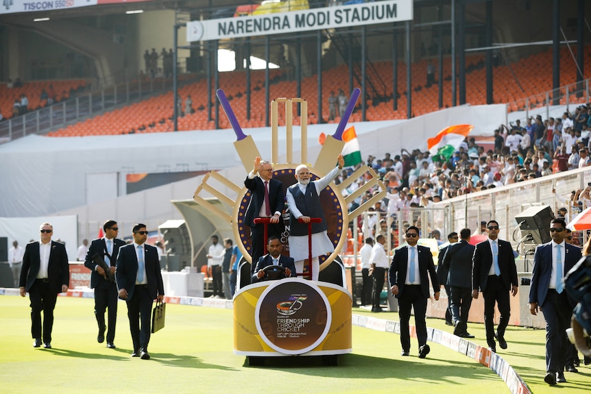 Anthony Albanese and Narendra Modi stand on a chariot with cricket bats on a cricket pitch surroudned by security.