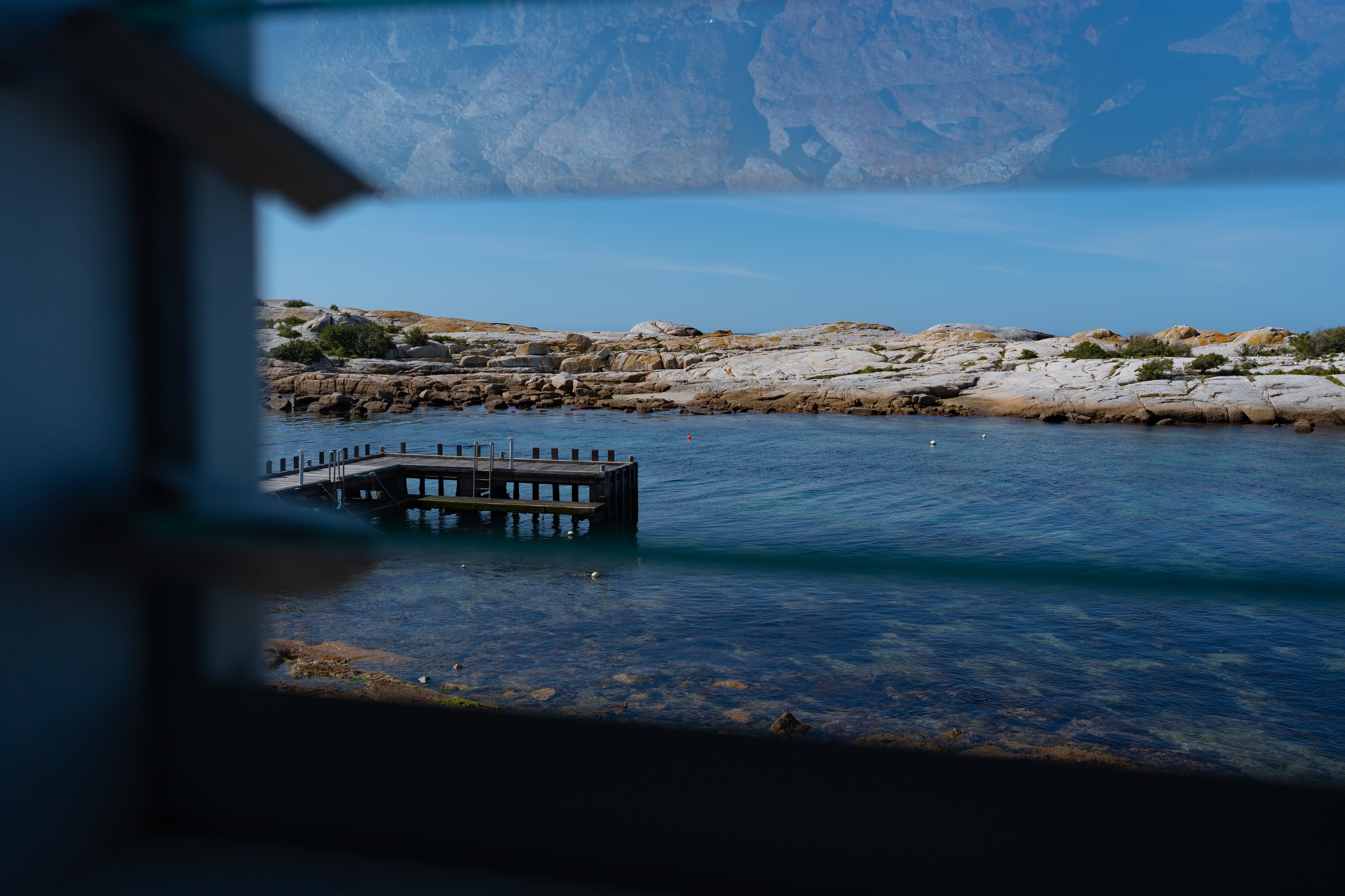 An old oyster hatchery sitting directly on the ocean on a clear day, there are rocks with orange lichen and a distillery