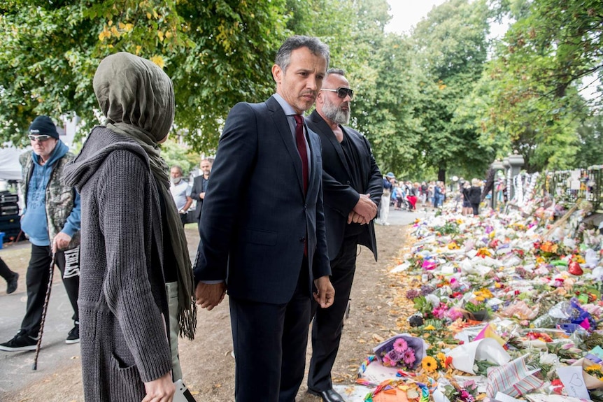 A man holds hands with his wife while looking at bunches of flowers at a memorial