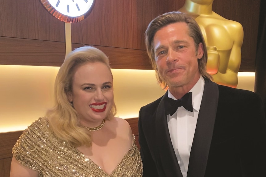 Rebel Wilson in a gold glittery formal dress with red lipstick and jewels standing next to Brad Pitt in a tux, near statue
