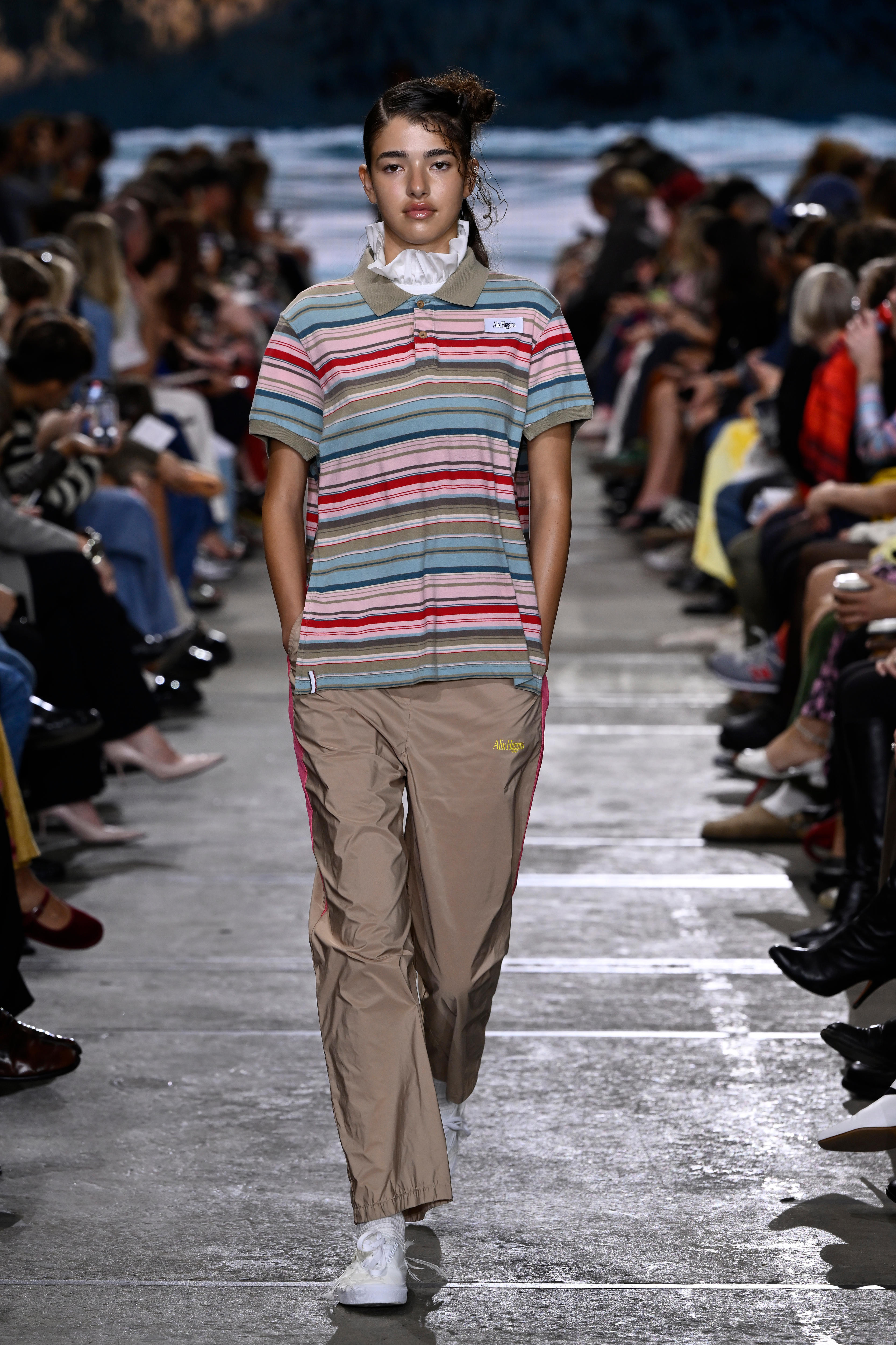 A model wears brown trakcpants and a multi-coloured polo shirt on the runway.