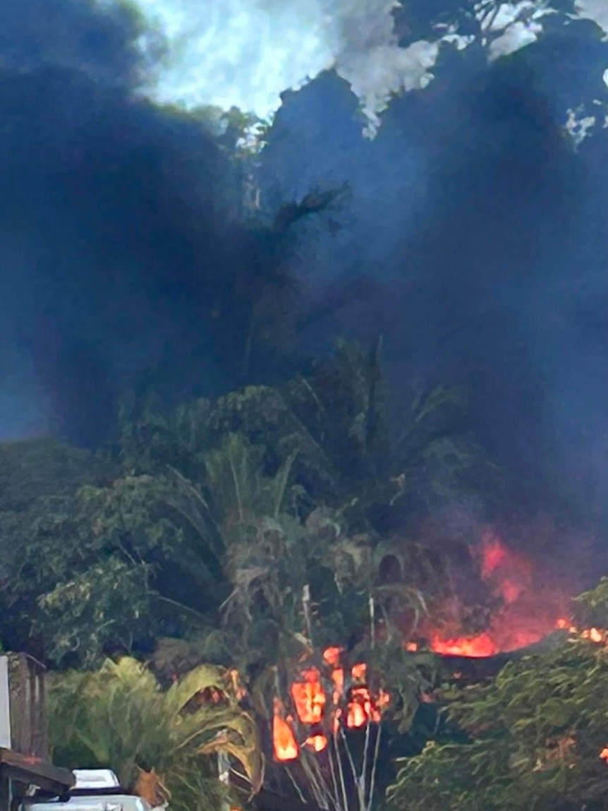 A fire burns behind palm trees with thick black smoke covering the sky