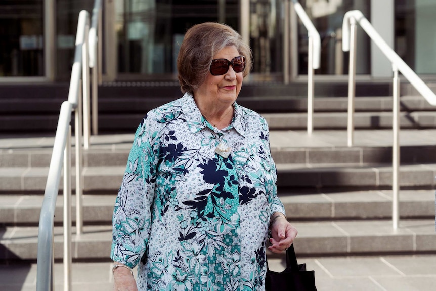 An older woman wearing a patterned shirt in front of stairs.