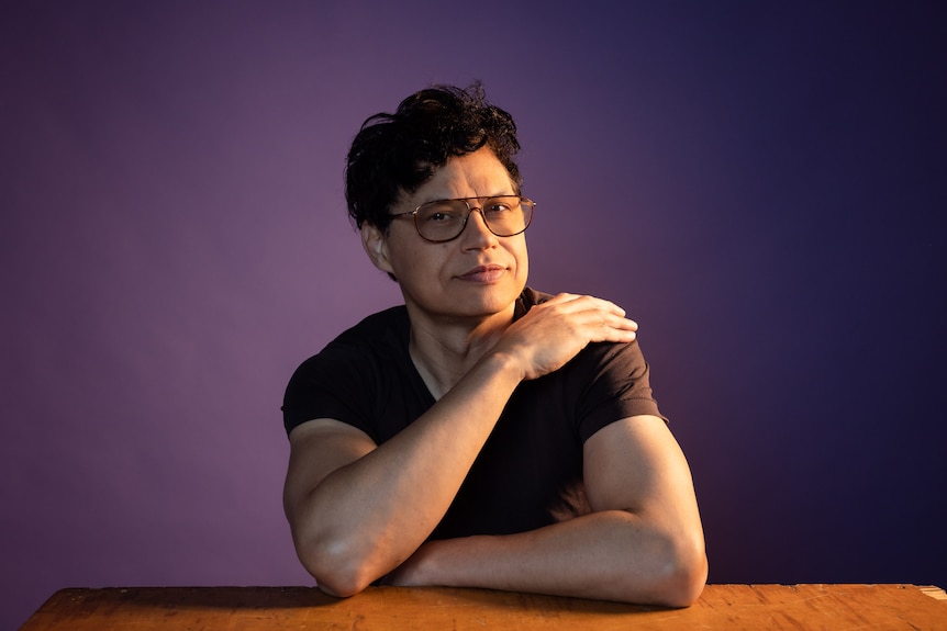 An Indigenous Australian man with curly dark hair and glasses poses for a photo, sitting at a table against a purple backdrop.
