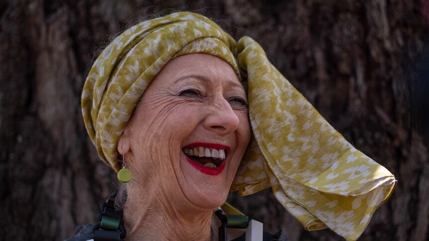 a woman with a yellow head scarf and blocky necklace laughs and smiles widely
