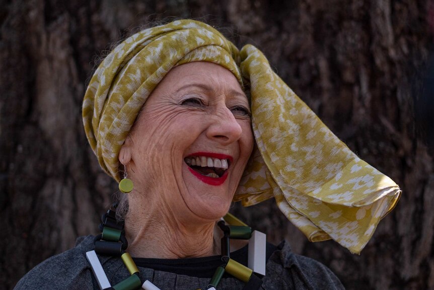 a woman with a yellow head scarf and blocky necklace laughs and smiles widely