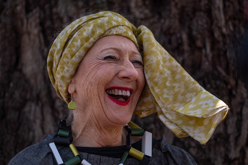 a woman with a yellow head scarf and blocky necklace laughs and smiles widely