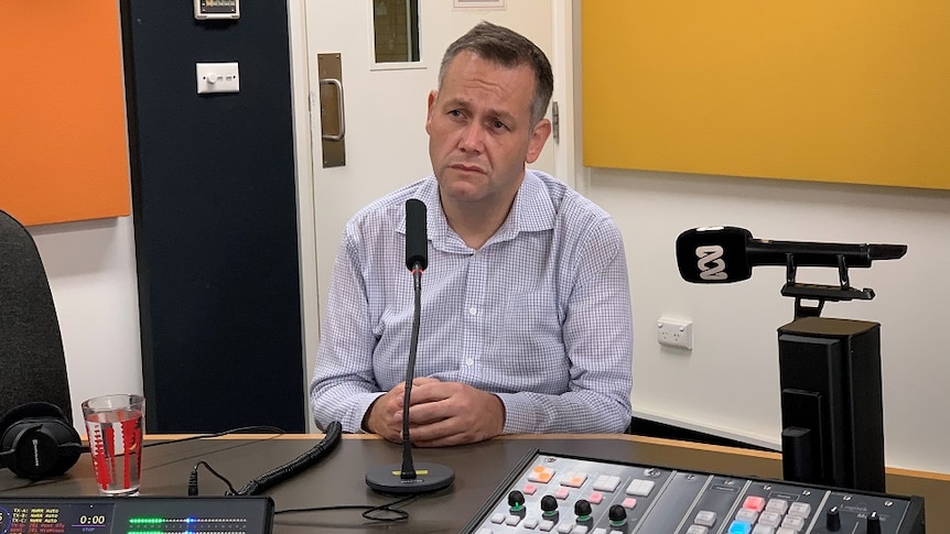 A middle-aged man with short hair, wearing a light-coloured shit, sits in a radio studio with a concerned look on his face.