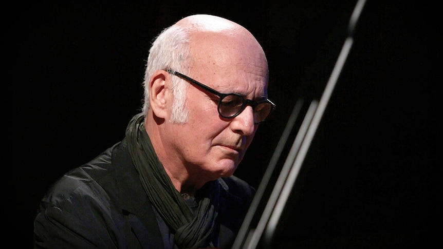 Composer Ludovico Einaudi plays the grand piano. He wears black-framed glasses and has a contemplative look on his face.