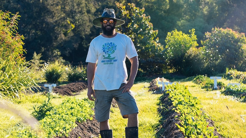 A man wearing gumboots and a broad-brimmed hat stands among rows of produce in a market garden.