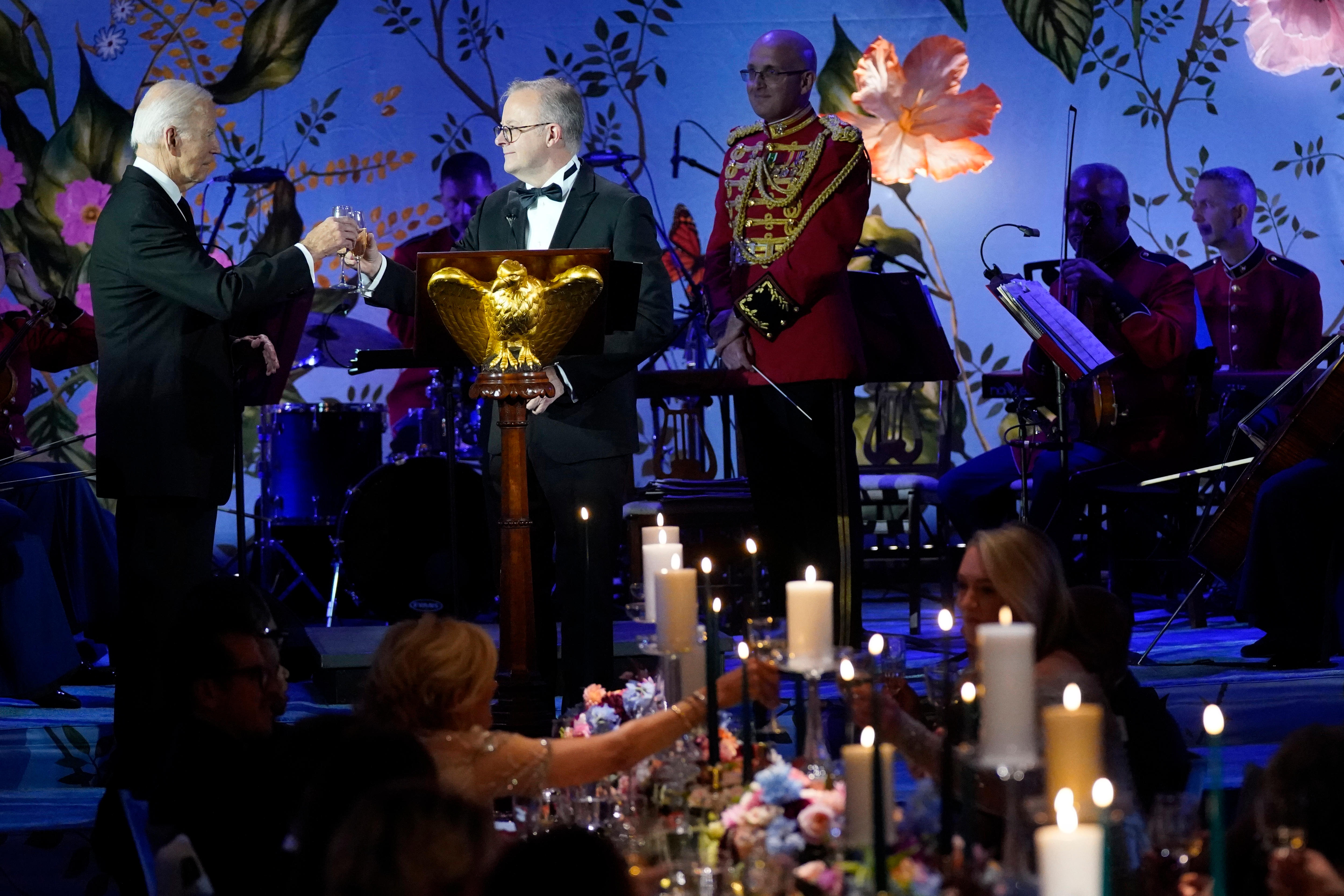 Anthony Albanese and Joe Biden clink champagne glasses on stage in a room with purple lighting and colourful decorations.
