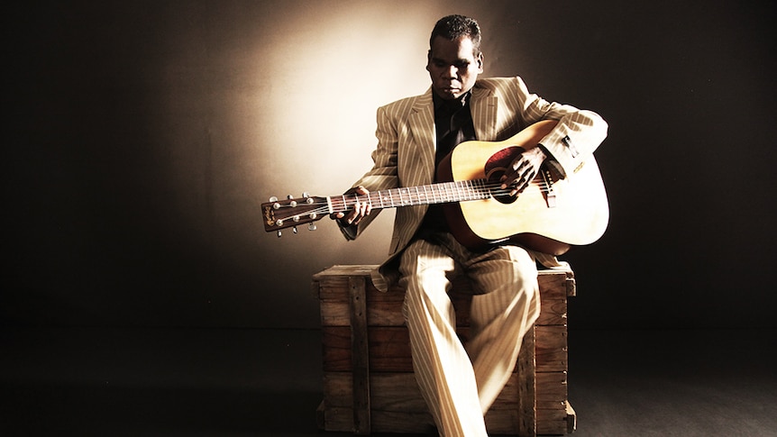 Gurrumul sits on a wooden crate in a tan suit with white stripes playing the guitar.