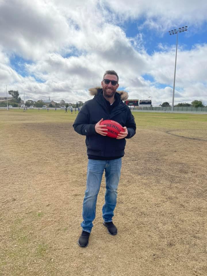 A man wearing blue jeans, a navy jacket and wearing black sunglasses holds a red AFL football smiling. 