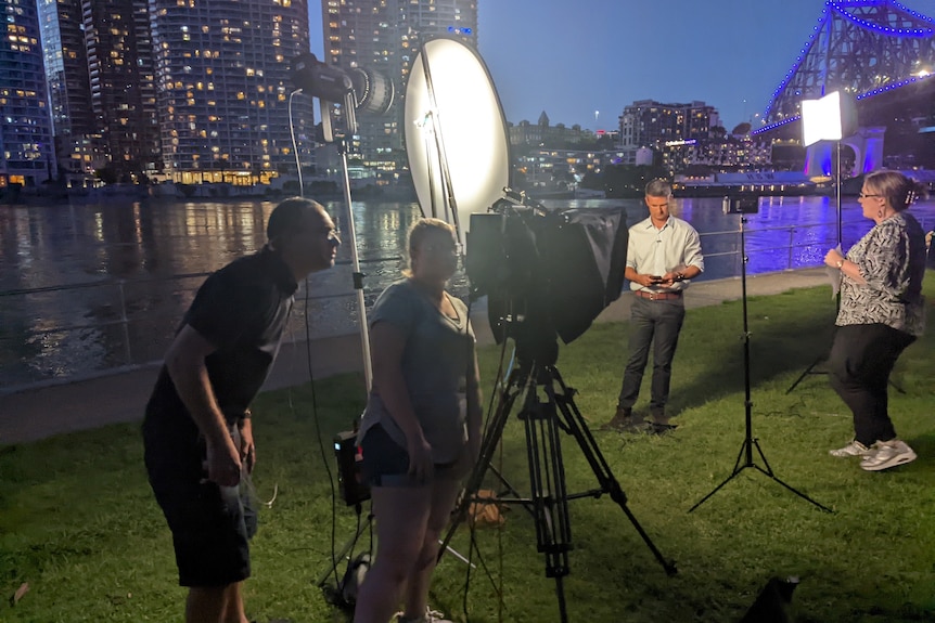 Camera crew set up on riverbank at night with presenter preparing to go to air, surrounded by lights and reflector board.