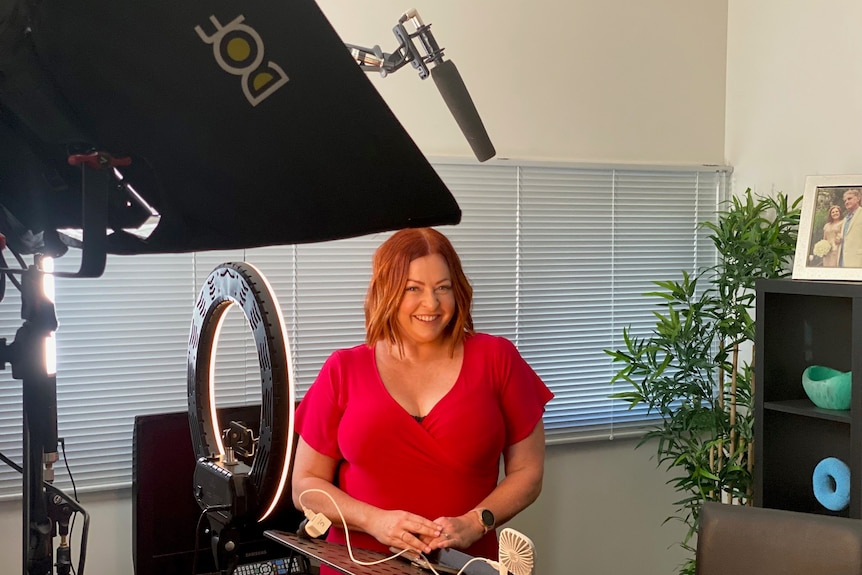 A women in a bright red dress smiling at the camera surrounded by TV lights in a home office