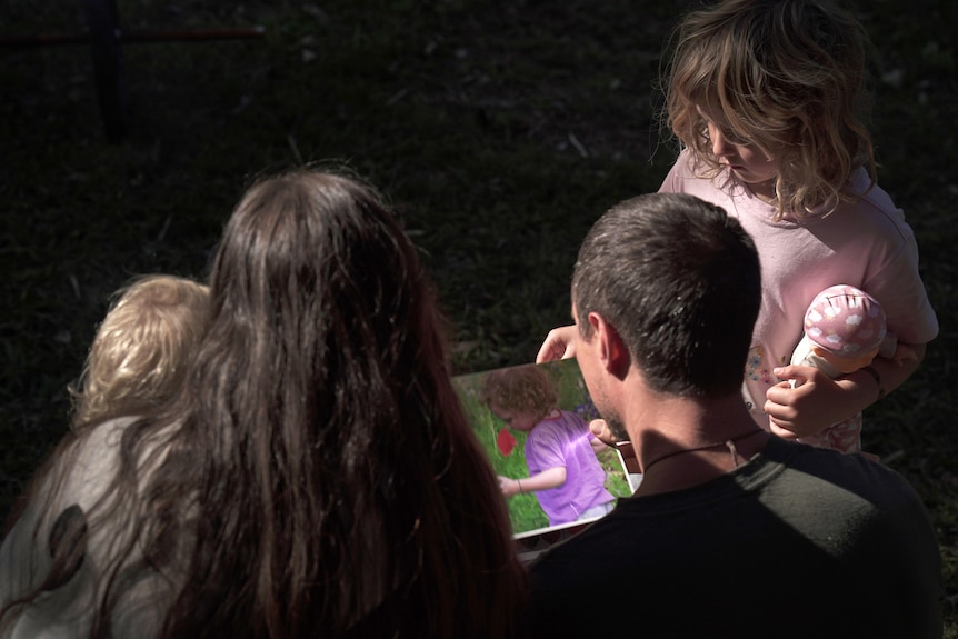 A man and woman and their two children from behind look at a photo album which includes a photo of a little girl