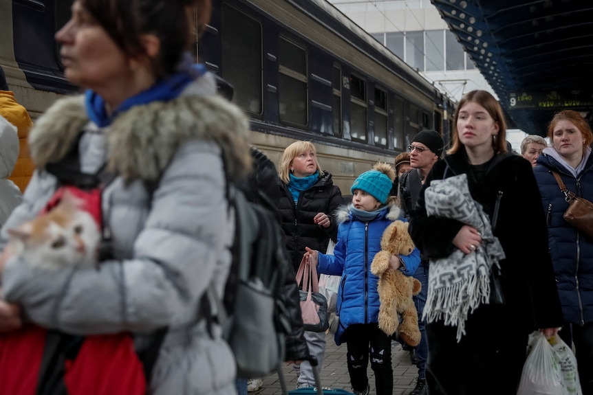 A group of people wearing winter clothes and holding luggage and pets walk along a train station platform, next to a train.