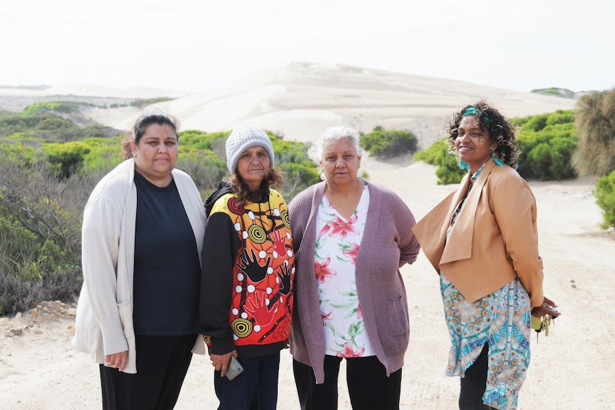 Four indigenous women standing on a road in front of a sandhill and green shrubs on side of road, not looking happy