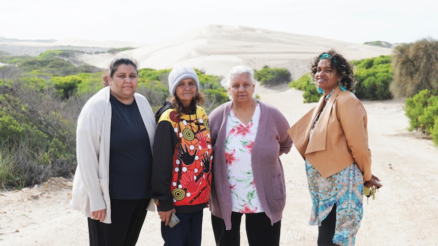 Four indigenous women standing on a road in front of a sandhill and green shrubs on side of road, not looking happy