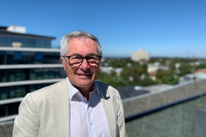 Man in glasses stands on roof top or balcony with cityscape behind him.