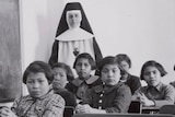 A black and white photograph of Canadian First Nations school children with a Nun in a traditional habit standing behind them.