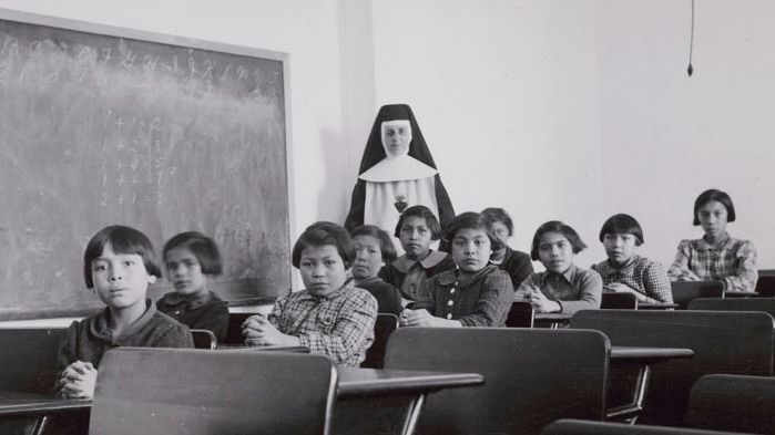 A black and white photograph of Canadian First Nations school children with a Nun in a traditional habit standing behind them.