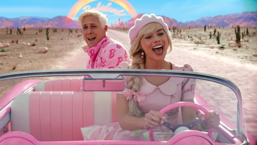 A blonde white man wears a pink shirt, while riding in the back of a pink car driven by a white blonde woman in a pink outfit.