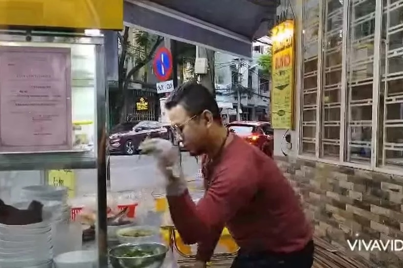 A still from a video of a Vietnamese man ith red shirt sprinkling herbs onto a soup.