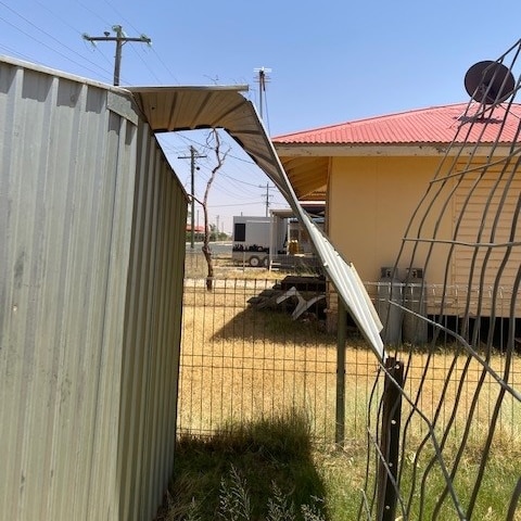 A tin shed with its roof torn off in front of a yellow house, damaged by storms.