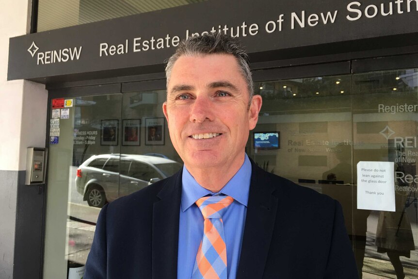 A man in a suit and a tie smiles outside a building with a sign "Real Estate Institute of NSW".