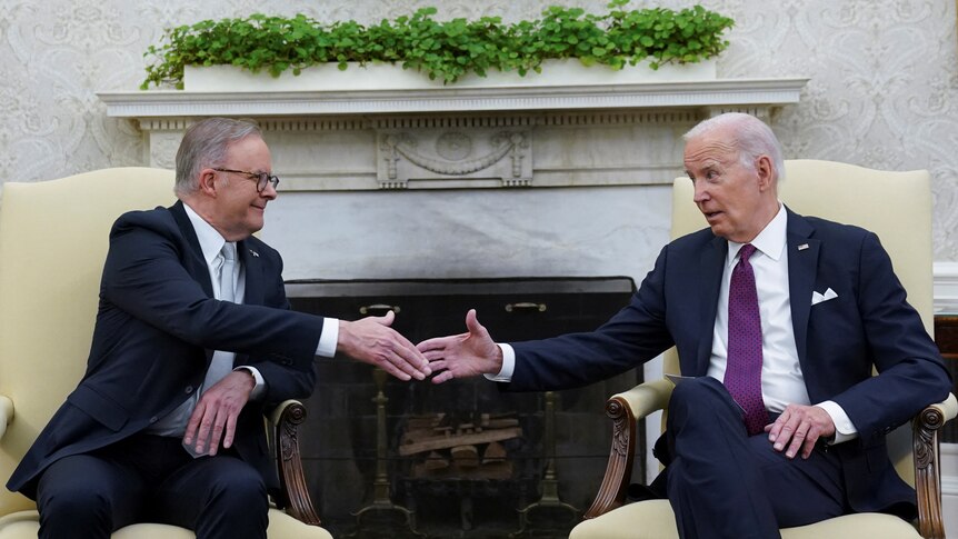 Anthony Albanese and Joe Biden shake hands while sitting in cream coloured chairs in front of an unlit fireplace.