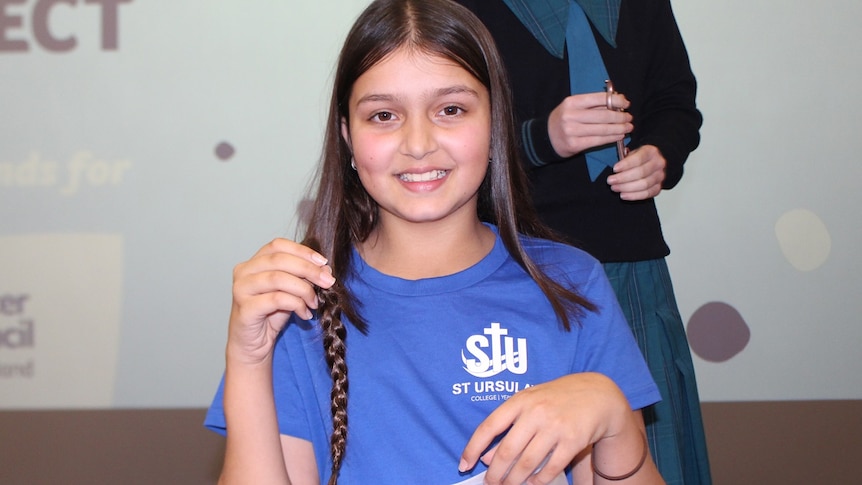 Priya sitting in a chair, wearing a blue t-shirt smiling, hold her ponytail that has been cut off.