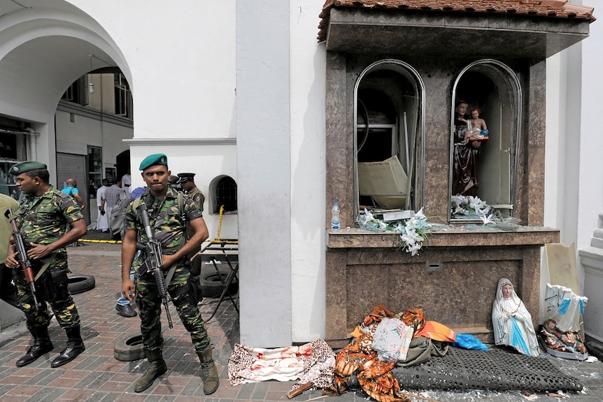 Soldiers guard a destroyed religious shrine, with a statue split in half among debris on the ground
