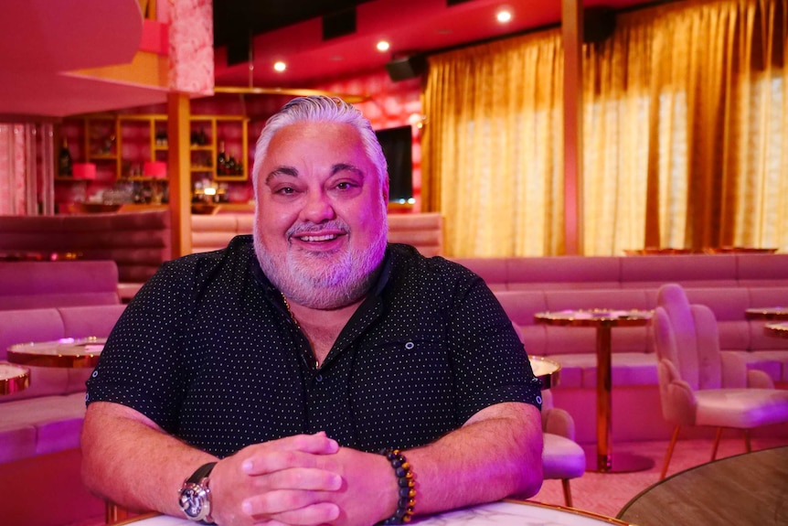 A man with short, white hair and a beard sitting in a venue, smiling.