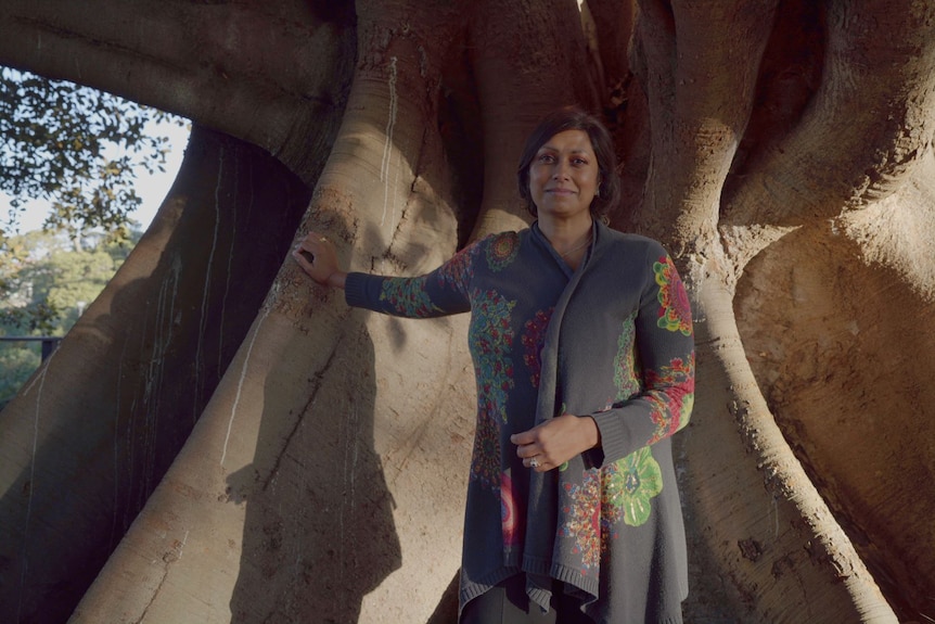 Indira Naidoo stands next to the giant trunk of her tree, wears a long gray cardigan, bright flower print on it, smiles sadly.