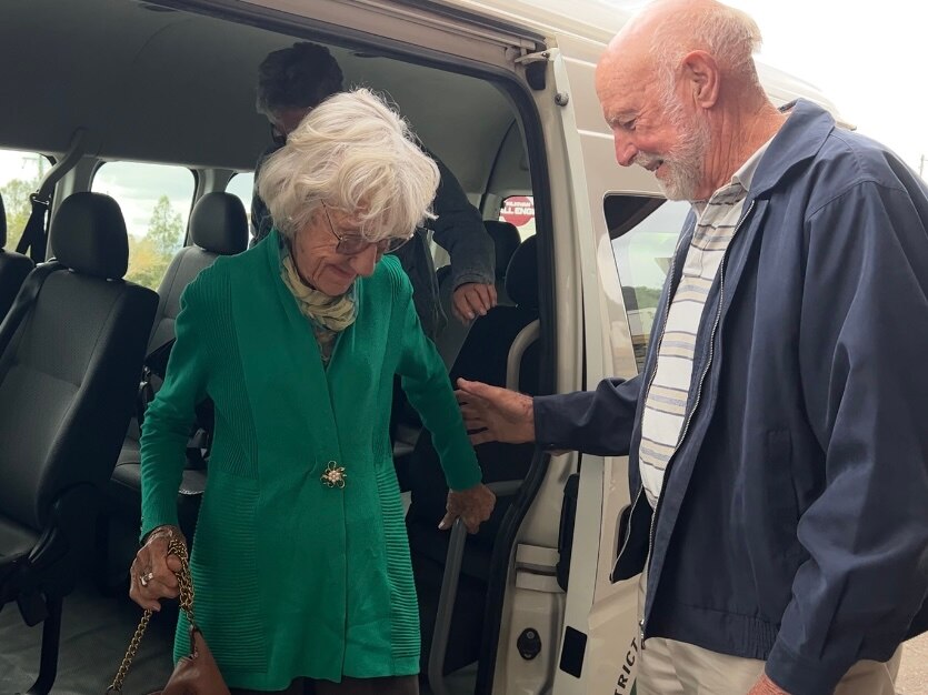 An older balding man helps an elderly woman dressed in green and scarf around her neck off a bus.